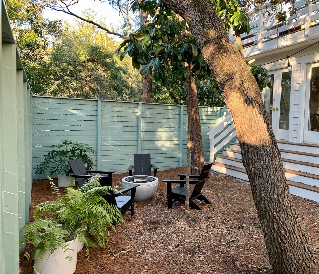 Green fenced side yard with black Adirondack chairs around fire pit